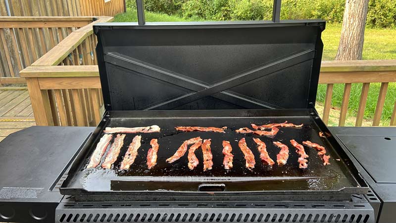 bacon cooking unevenly across the flat top of the camp chef gridiron 36 with a fan on one side